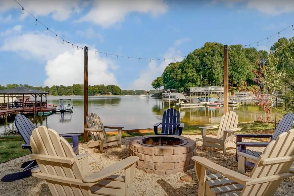 The image showcases a picturesque lakefront setting featuring a glowing fire pit surrounded by inviting Adirondack chairs, all beneath twinkling string lights. Beyond, a tranquil lake shimmers at sunset with a private dock extending out over the water. The scene exudes warmth and togetherness, inviting guests to gather, relax, and enjoy the natural beauty.
