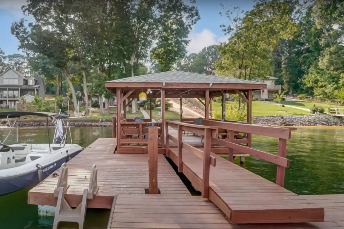 A peaceful lakeside scene featuring a wooden dock extending into the water, a charming gazebo, a moored boat, and lush greenery surrounding the shoreline. The soft light suggests either a tranquil sunrise or sunset, enhancing the inviting atmosphere for outdoor activities.