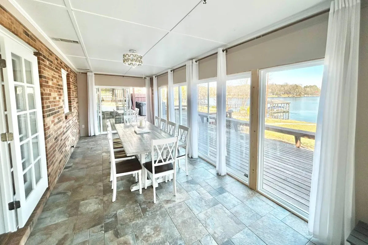 A spacious interior showcases a long dining table set for a gathering, with large windows revealing stunning panoramic lake views. The brick accent wall adds warmth, while the arrangement of chairs hints at group meals and lively conversation. Soft evening light streams in, inviting celebrations and togetherness.