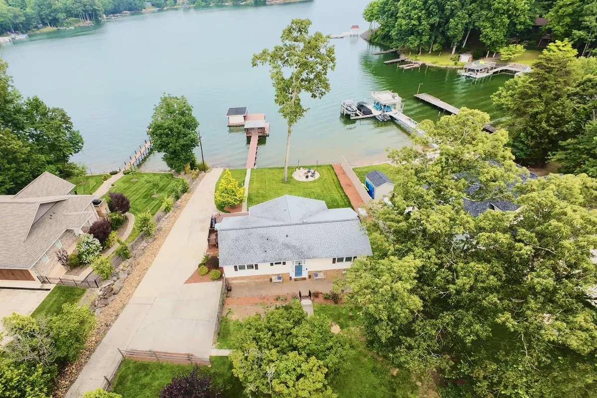 This exterior image displays the lakeside property with the main house, a green lawn, the tranquil lake, and dock structures extending into the water. The scene is bathed in natural light, suggesting both playful and relaxing outdoor opportunities.