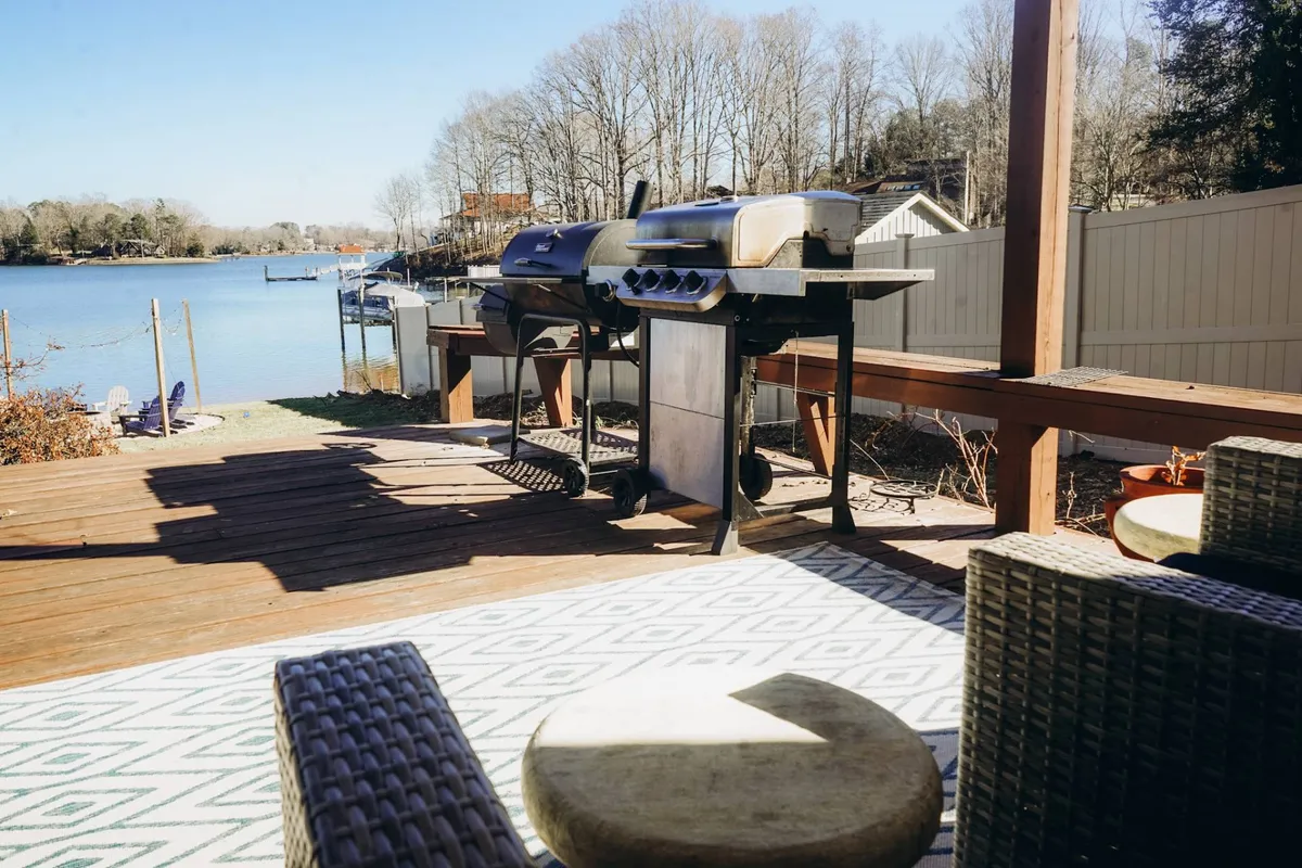 A beautiful exterior deck area featuring a grill, outdoor seating, and a clear view of the tranquil lake. The scene is set for barbecuing and al fresco dining, with spaces ideal for social gatherings and relaxing by the water. Sunset lighting adds a warm, inviting glow.