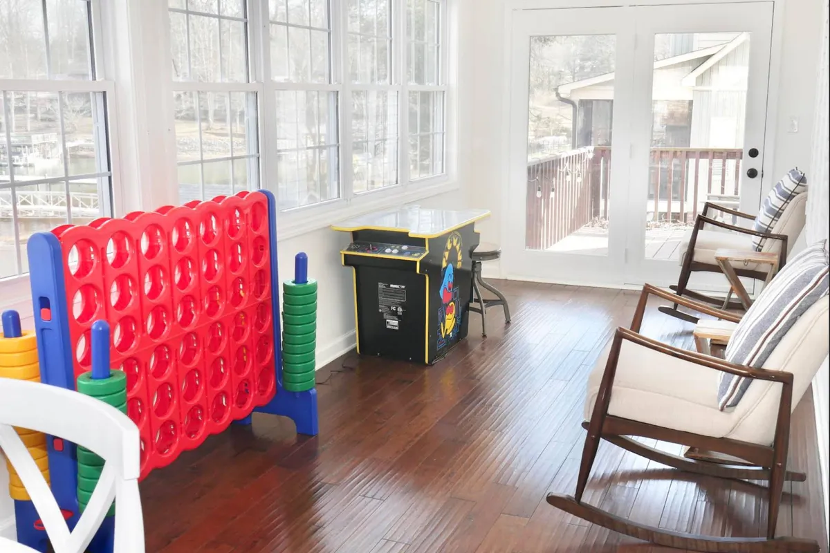 This lively sunroom features a large red Connect Four game, an arcade game machine, and comfortable rocking chairs set against expansive windows that flood the space with natural light. The arrangement is inviting and hints at group fun, relaxation, and friendly competition.