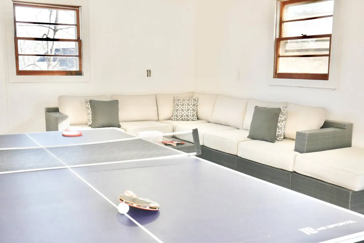 A spacious game area with a ping pong table as the centerpiece, joined by a comfortable sectional sofa adorned with colorful pillows. The scene suggests a relaxed atmosphere where guests can alternate between spirited matches and lounging with friends.