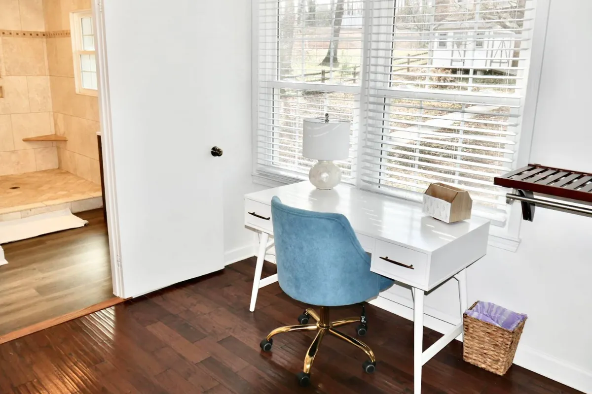 A functional suite corner with a desk and blue chair positioned by a window, allowing natural daylight to illuminate the workspace. In the background, a glimpse of a private bathroom area is visible, reinforcing the sense of a multi-purpose master suite.