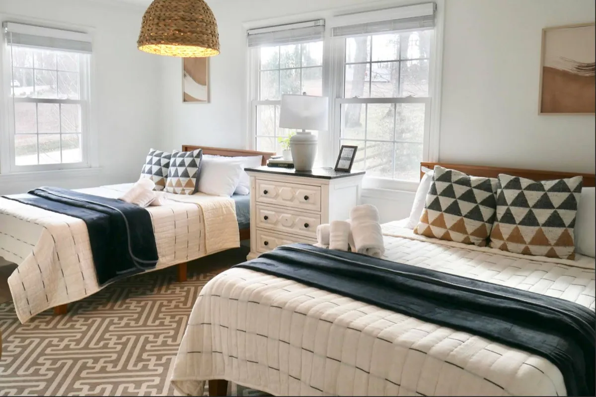 A welcoming bedroom featuring two beds with decorative pillows, a woven pendant light, a nightstand with a lamp, and plenty of daylight streaming in. The space feels cozy and is ideal for families or groups.
