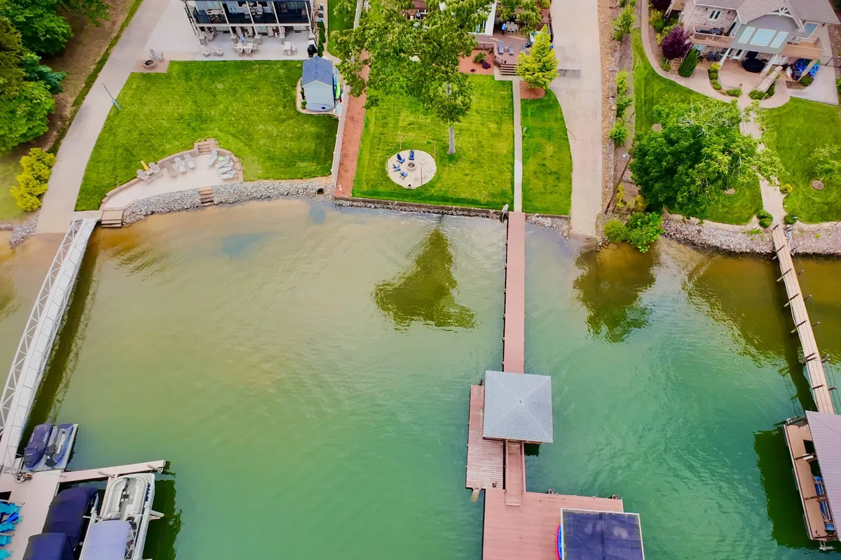 A wide view of the dock leading out onto the lake, with an adjacent lawn and a cozy seating area on the waterfront. Other houses are visible across the water, hinting at a friendly lakefront community and ample outdoor space perfect for picnics, water activities, or letting pets play.