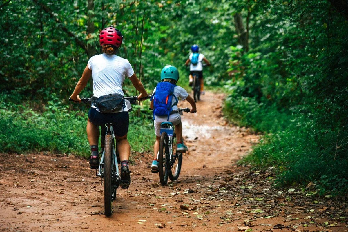 An exterior action shot of cyclists riding along a winding dirt path surrounded by lush greenery. The scene is energetic and joyful, emphasizing outdoor activity and exploration.